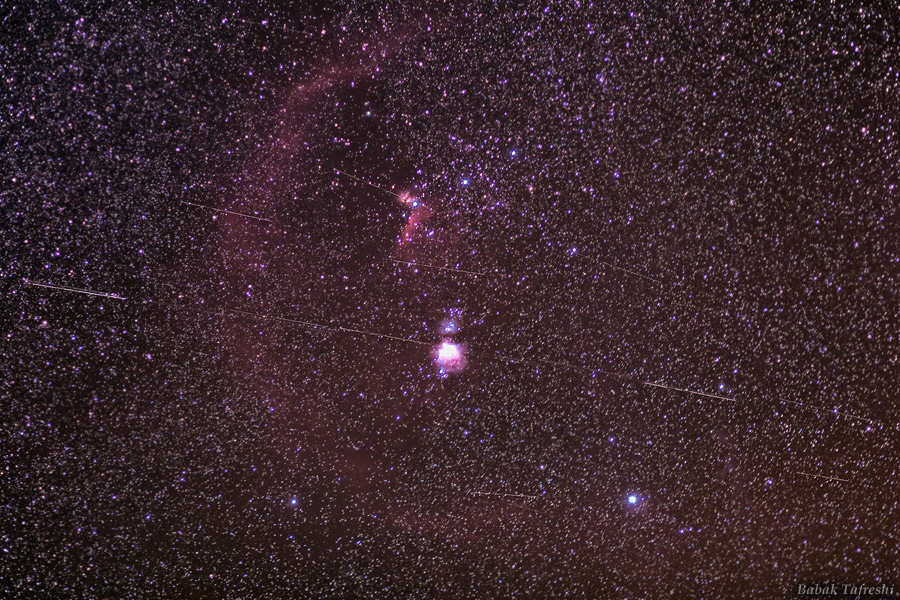 Mitten im Bild ist der Orionnebel, darüber die drei Gürtelsterne. Im Hintergrund sind viele Sterne verteilt. Quer über dem Orionnebel verlaufen mehrere Lichtspuren von geostationären Satelliten.