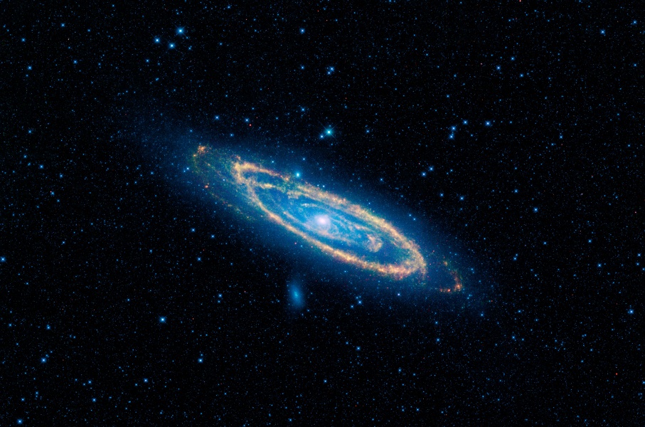 Im Bild schwebt eine schräg von oben sichtbare Spiralgalaxie, sie schimmert blau mit hellem, rosafarbenem Zentrum und sehr markanten hellorange gefärbten Staubbahnen entlang der Spiralarme. Es ist die Andromeda-Galaxie in Infrarot, die hier kaum erkennbar ist.
