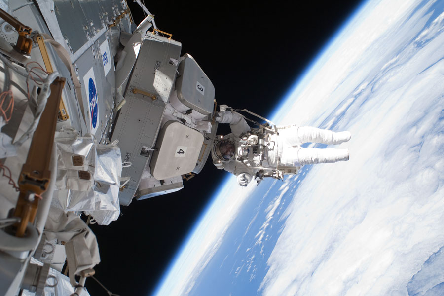 Ein Astronaut hängt außen an der Cupola der Internationalen Raumstation. Im Hintergrund leuchtet rechts unten die Erde, sie füllt fast das halbe Bildfeld.