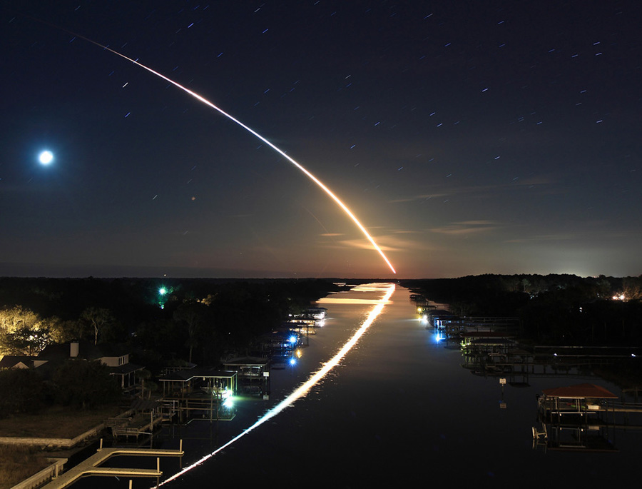 Über einem Gewässer steigt eine Rakete nach links auf, ihre Leuchtspur spiegelt sich im Wasser. Links am Bildrand steht der Mond.