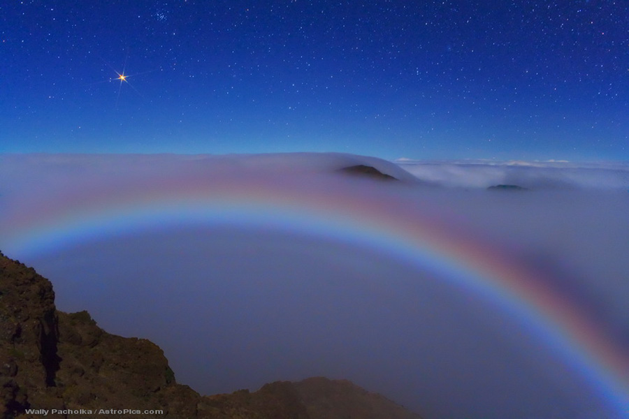 Hinter einer Felsklippe breitet sich ein Nebelmeer aus, in dem ein regenbogenfarbiger Nebelbogen leuchtet. Darüber leuchtet am klaren Himmel ein Licht, der Planet Mars.