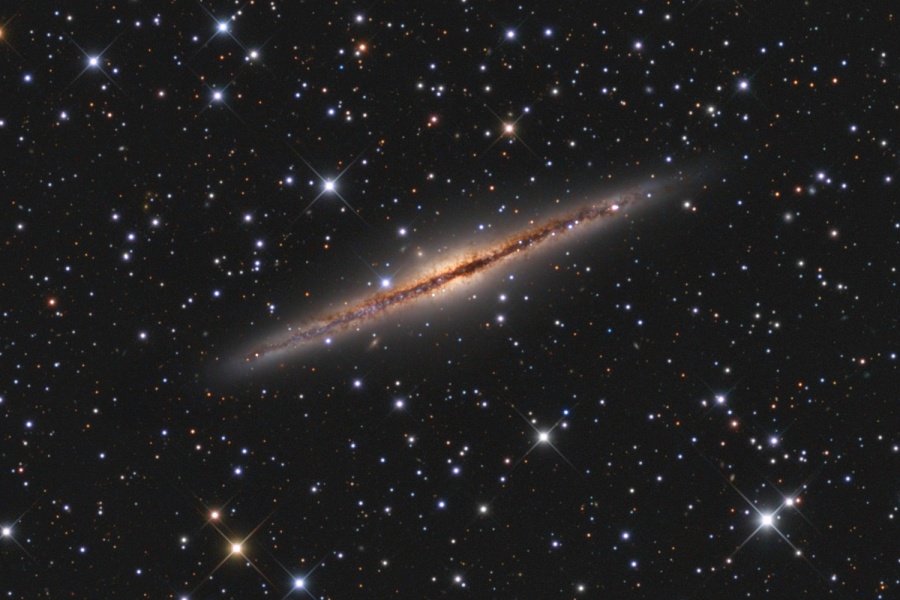 Schräg im Bild liegt eine von der Kante sichtbare Spiralgalaxie. Durch die Mitte läuft eine markante dunkle Staubbahn. Im Hintergrund sind unterschiedlich helle Sterne lose verteilt.