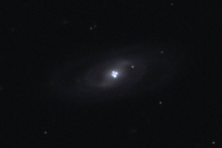 Nitten im Bild leuchten vier eng beisammen stehende Lichtflecken, umgeben von einem blassen galaxienförmigen Nebel. Der Rest des Bildes ist dunkel mit wenigen sehr blassen Lichtpunkten.