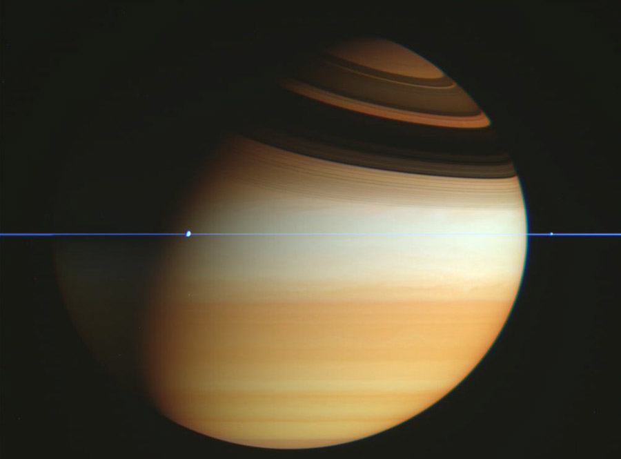 Der dreiviertel beleuchtete Saturn füllt das Bild, die Wolken verlaufen waagrecht, oben sind die Schatten der Ringe, die waagrecht als dünne blaue Linie durchs Bild verlaufen. Im Ring zeichnen sich zwei Monde ab.