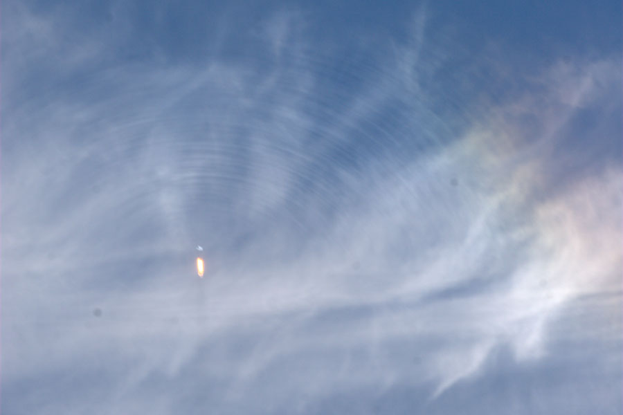 Am blauen Himmel sind Cirruswolken, in der Mitte eine startende Rakete, von der Dichtewellen ausgehen. Rechts oben ist ein Halo.