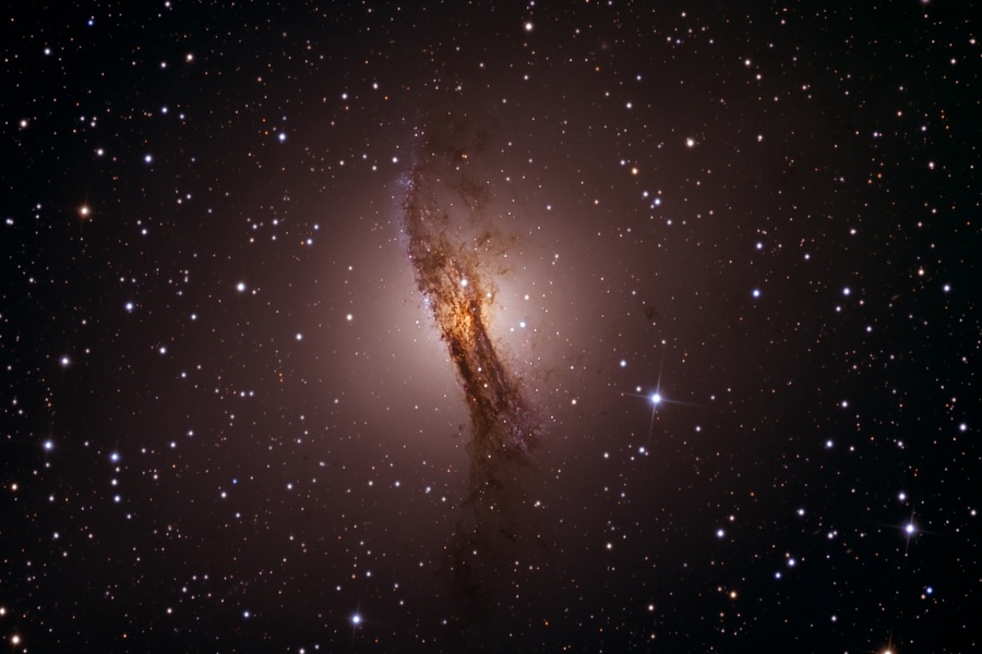 In der Bildmitte leuchtet ein nebeliges rundes Objekt inmitten von Sternen, über die Mitte verläuft ein stark strukturiertes dunkles Staubband.
