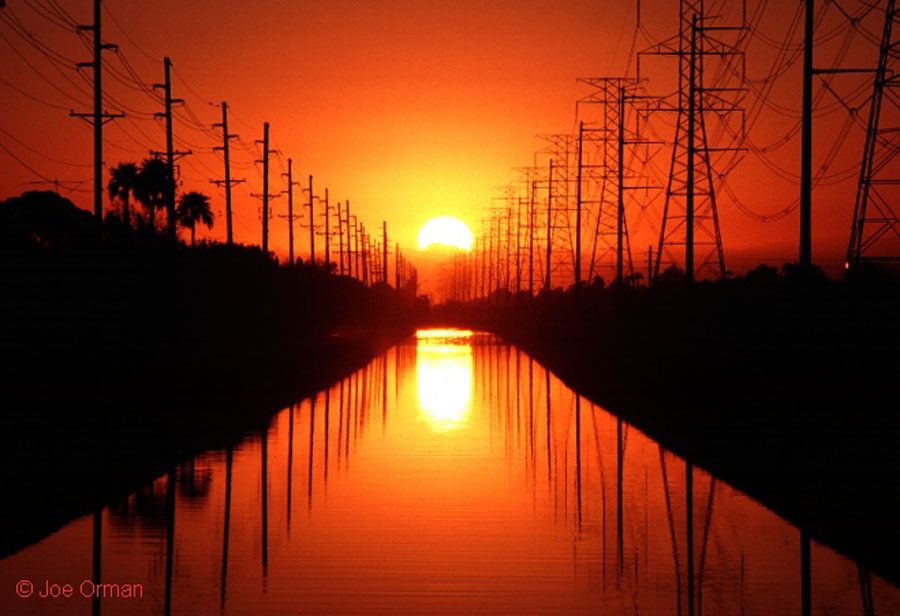 Am Ende eines langen Kanals mit Strommasten links und rechts geht die Sonne unter, der Sonnenuntergang spiegelt sich im ruhigen Gewösser. Der Himmel leuchtet orangefarben, die Landschaft links und rechts vom Kanal ist dunkel.