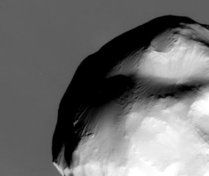 Das Schwarzweißbild zeigt einen runden Himmelskörper mit glatten Oberflächen, die wie Hänge wirken. Dahinter ist die Oberfläche von Saturn.