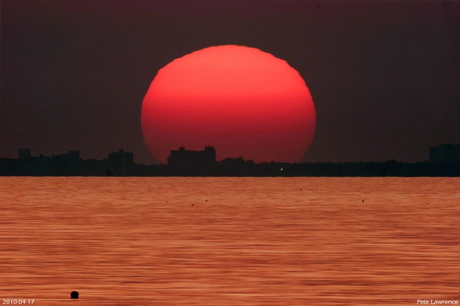 Über einem Gewässer geht eine leuchtendrote Sonne unter. Am Horizont ist eine dunkle Skyline.