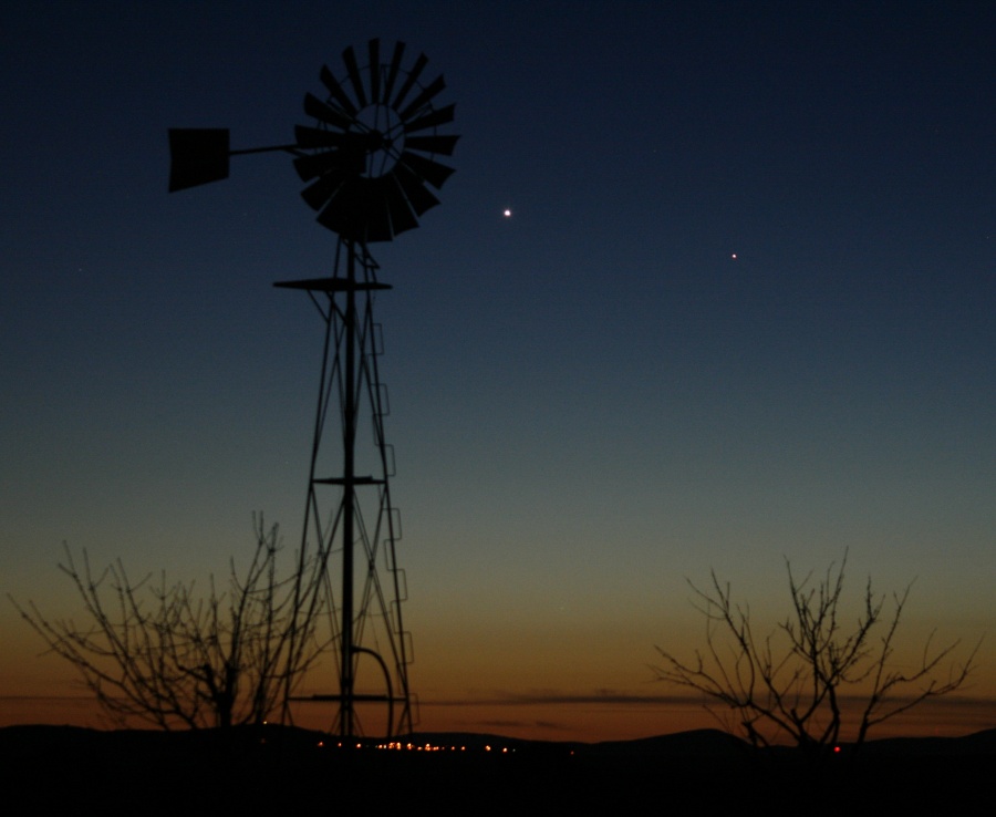 Am Horizont mit Büschen links und rechts und dem Gestell eines Windrades ist der Himmel dunkelrot befärbt und geht über dunkelgelb in Blau und Schwarz über. Am Himmel leuchtet die Venus und rechts davon etwas tiefer Merkur.