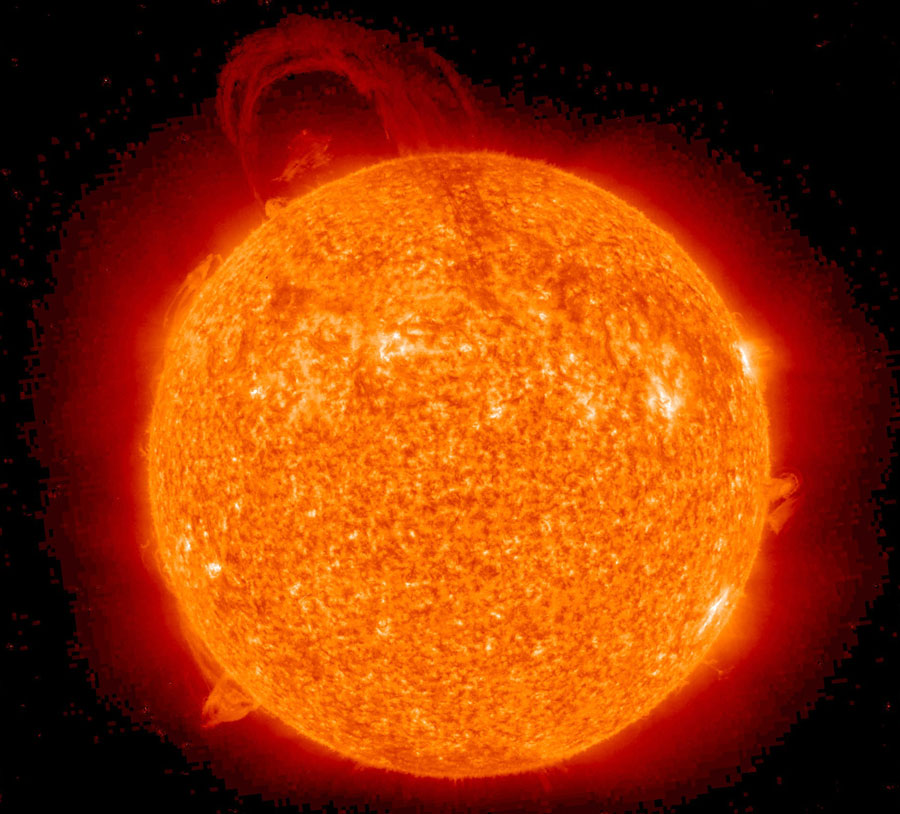 Ein vollständiger Sonnenball schwebt im Raum. Die Sonne leuchtet rot und orangefarben mit einigen hellen Flecken, sie ist von dunkelroten Nebeln umgeben, an einigen Stellen steigen Protuberanzen hoch, oben ist eine riesige Sonnenfackel.