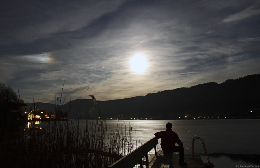 Ein zugefrorener See wird von Mondlicht beschienen. Am leicht bewölkten Himmel ist ein Nebenmond erkennbar.