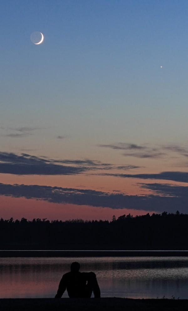 Vor einem See ist Eine Gestalt als Silhouette erkennbar, dahinter ein dunkler Wald. Der Himmel darüber verläuft von rosarot bis dunkelblau, unten sind Wolken, darüber leuchtet am klaren Himmel links der Sichelmond, rechts die Venus.