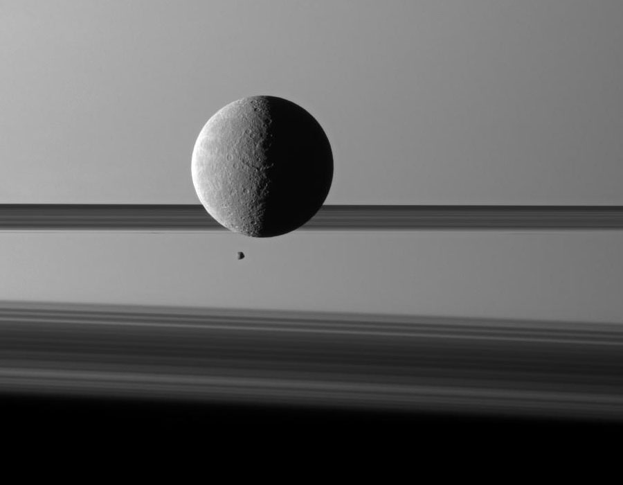 Hinter einem schwarzweißen Mond mit Kratern schwebt ein viel kleinerer zweiter Mond, dahinter werfen die Saturnringe Schatten auf die Oberfläche des Ringplaneten.