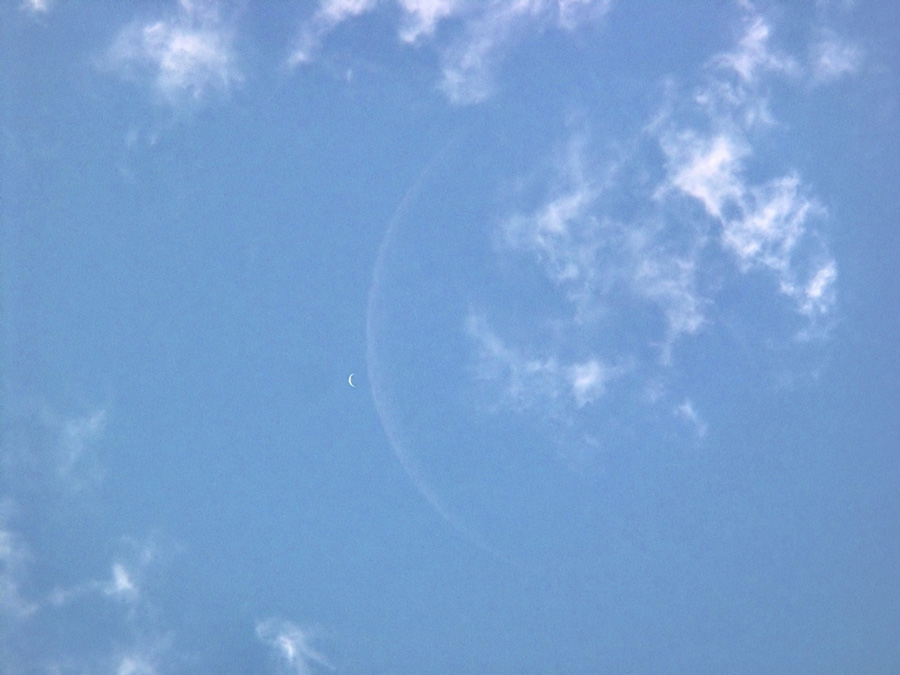 Am blauen Himmel schweben zarte Wolkenfetzen. Wenn man sehr genau hinsieht, erkennt man zwei Sicheln, die kleine kompakte, hellere Sichel ist die Venus, die blasse, viel größere Sichel ist der Mond.
