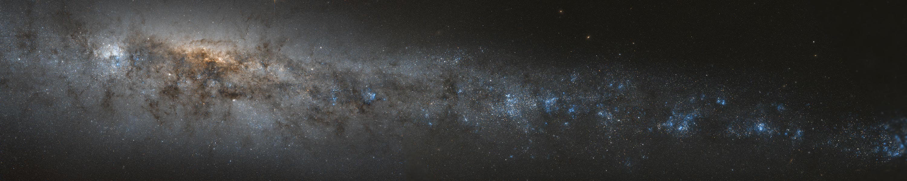 Das Panorama zeigt eine von der Seite sichtbare Galaxie mit vielen dunklen Staubwolken und blauen Sternbildungsregionen.
