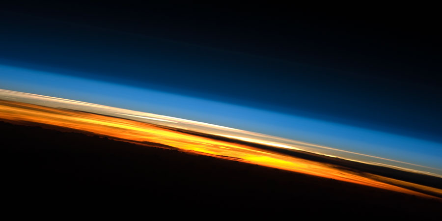 Hinter der dunklen Erde geht die Sonne unter. Unten ist ein orangeroter Streifen mit Wolken, darüber eine hellblaue Schicht der oberen Atmosphäre, die in die Schwärze des Weltraums übergeht.