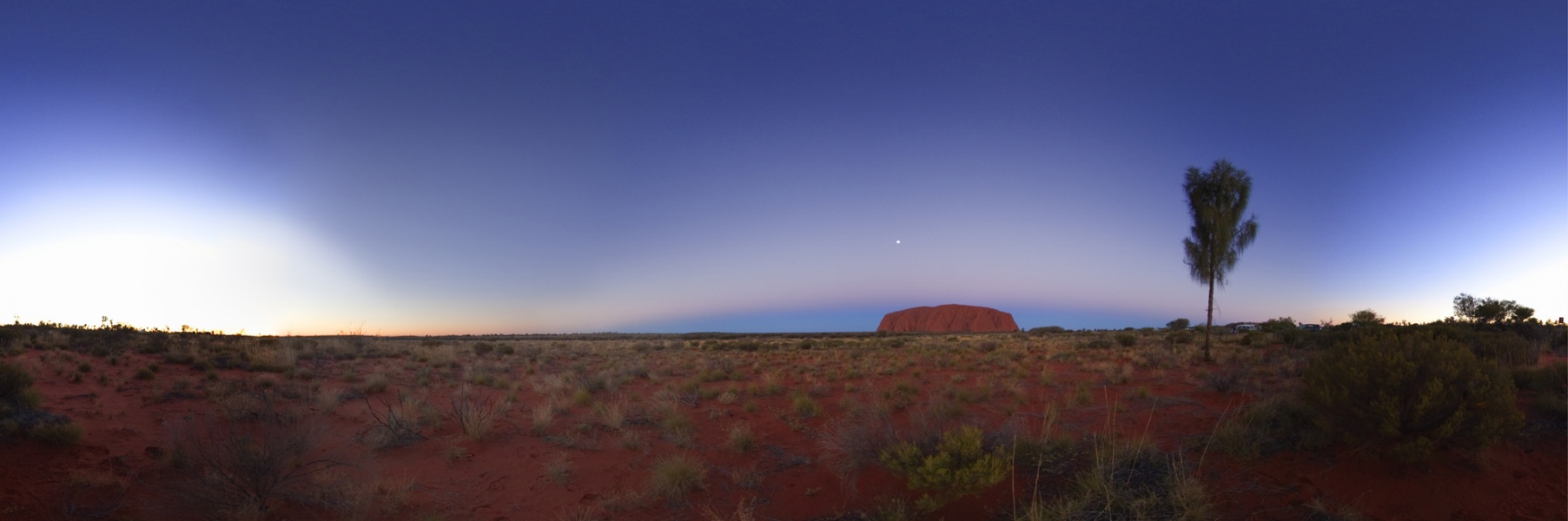 Das Panorama zeigt die Landschaft um den Uluru in Australien. Links ist der helle Himmelsteil, wo die Sonne untergegangen ist, rechts neben der Mitte ragt Ayers Rock auf, dahinter steigt der Erdschatten auf. Der Himmel ist glasklar.