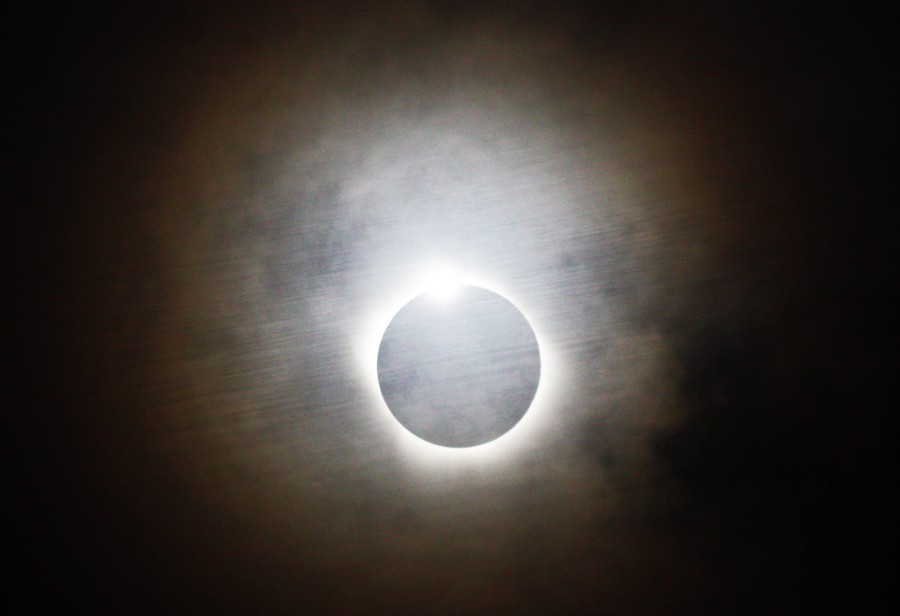 Die Sonne hst hinter Wolken und hinter dem Mond versteckt. Am oberen Mondrand blitzt die Sonne hervor, rund um den Mondrand leuchtet die Corona, die Wolken davor sind ebenfalls hell mit einem roten Rand in einiger Entfernung zur Sonne.