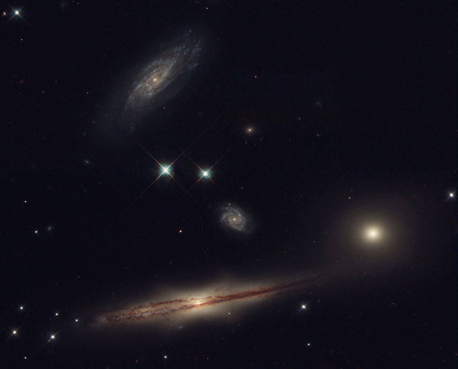 Mehrere Galaxien sin dim Bild verteilt, oben eine zerfledderte Spiralgalaxie, unter zwei hellen Sternen eine kleine, von oben sichtbare Spiralgalaxie und rechts eine kleine elliptische Galaxie, unten eine flache Spiralgalaxie, die von der Kante sichtbar ist, mit einem markanten Staubwulst in der Mitte.