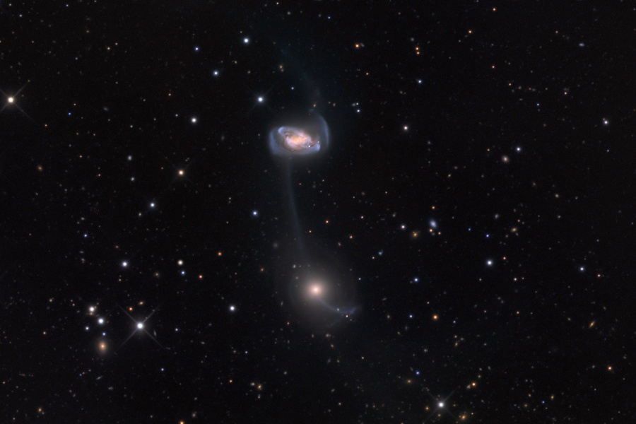 Im Bild mit wenigen Sternen sind in der Mitte zwei Galaxien übereinander, die untere wirkt strukturlos mit hellem Kern und Nebel rundherum, die obere hat verzerrte Strukturen.