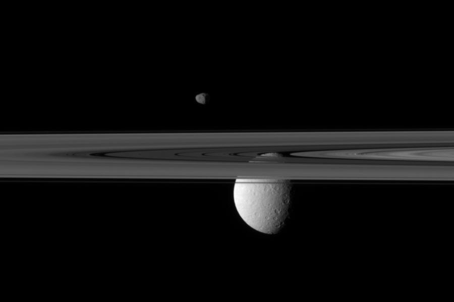 Unter den Ringen Saturns hängt ein grauweißer Mond mit vielen Kratern, über der Ringebene schwebt ein viel kleinerer Mond.