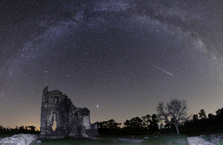 Über einer Ruine und einem Wald wölbt sich die Milchstraße. Rechts zischt ein Meteor über den Himmel.