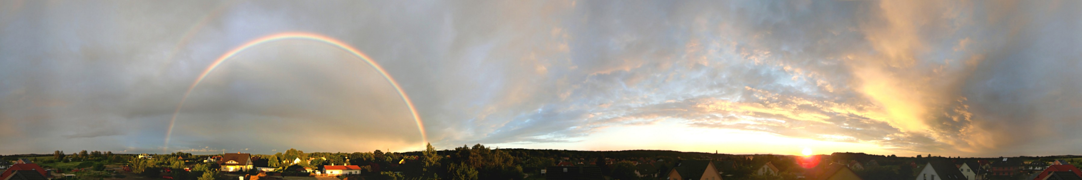 Im Panorama geht rechts die Sonne unter, links ist am Wolkehhimmel ein Regenbogen zu sehen.
