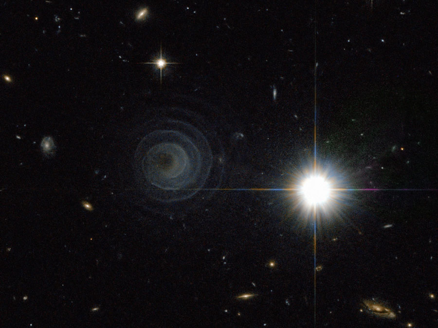 Im Bild leuchtet rechts ein heller, weißer Stern mit bunten Strahlen, rechts daneben ist eine zarte, sehr regelmäßig geformte Spirale um ein dunkles Inneres. Im Bild sind einige sehr ferne Galaxien verteilt.
