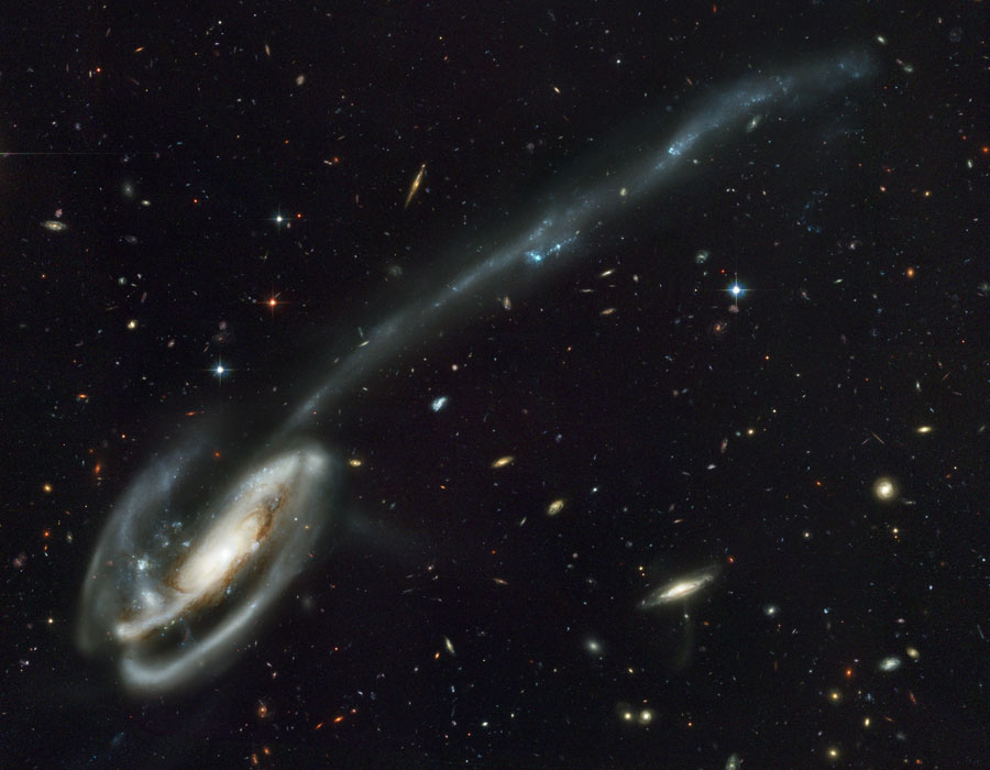 Links unten leuchtet das Zentrum einer Galaxie, die dreidimensional von Sternschleifen umwickelt ist. Ein Schweif aus Sternen ist nach rechts oben herausgezogen.