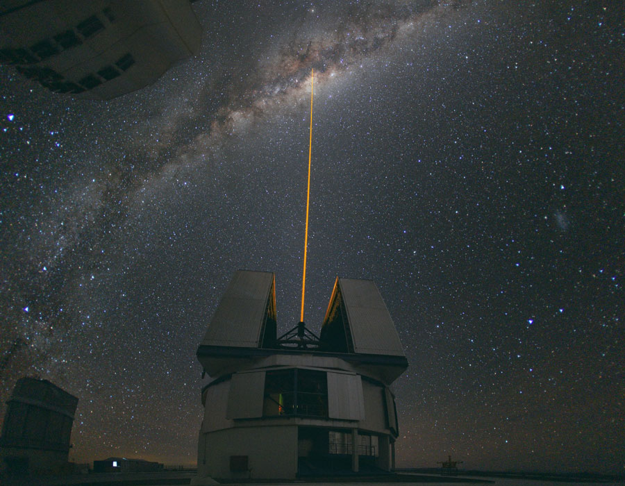 Mitten im Bild steht ein Observatorium mit geöffneter Kuppel. Nach oben schießt ein Laserstrahl ins Zentrum der Milchstraße, die von links unten aufsteigt. Der dunkle Himmel ist von Sternen übersät.