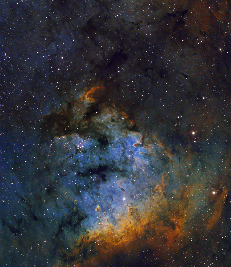 Im unteren Bildteil leuchtet ein matter blauer Nebel, oben wird er von dunklen Wolken begrenzt, unten von orangefarbenen Nebeln. Dazwischen sind dunkle Wolkenfetzen verteilt.