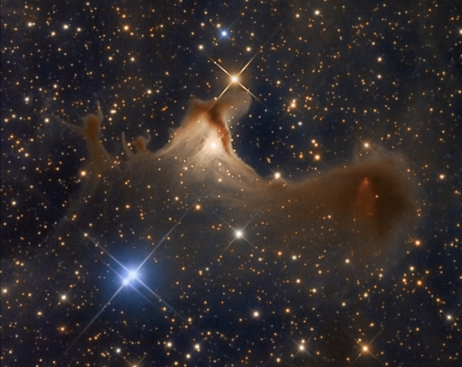 Vor bräunlich leuchtenden Sternen schwebt ein Nebelfetzen, der in der Mitte wie von hinten beleuchtet wirkt. Links unten strahlen zwei helle blaue Sterne dicht nebeneinander.