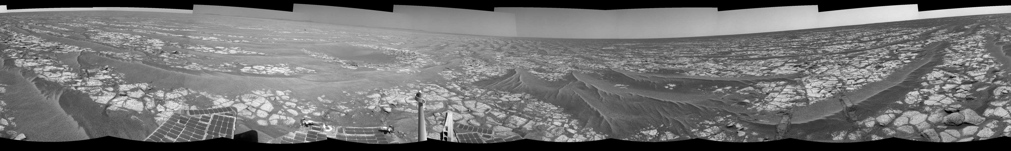 Das Panorama zeigt die Marsoberfläche mit hellem Gestein und dunklem Sand. Rechts sind Spuren des Rovers, am unteren Bildrand sind Solarpaneele und Instrumente des Rovers Opportunity.