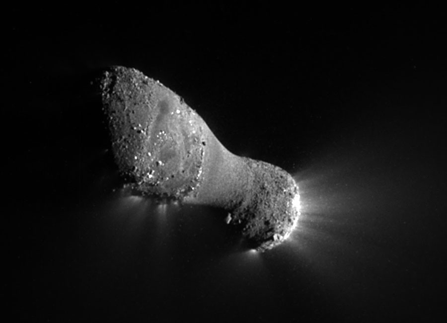 Der Komet im Bild wirkt länglich, in der Mitte ist er eingeschnürt und sehr glatt, die Enden sind mit Geröll übersät, am hinteren Ende strömen helle Strahlen aus.