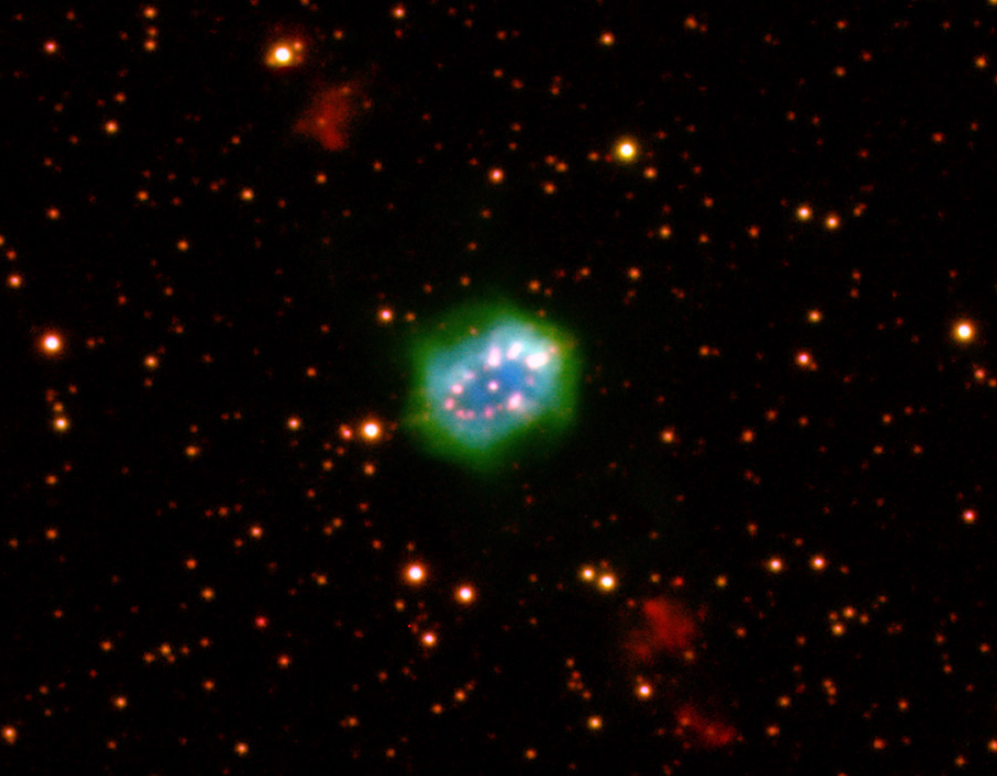 Mitten im Bild, das von matten Sternen mit roten Rändern befüllt ist, leuchtet ein Nebel, der an ein Perlenarmband erinnert, das von einem grün leuchtenden Nebel umgeben ist.