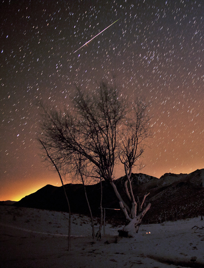In einer verschneiten Landschaft ragen Bäume auf, dahinter verläuft eine Bergkette. Ein Meteor zieht über einen orangefarbenen, von Strichspuren bedeckten Himmel.