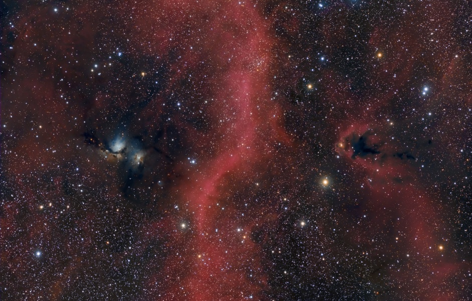 In der Mitte des Bildes verläuft eine leicht geschlängelte rote Nebelwulst. Links daneben sind zwei kleine weißliche Reflexionsnebel. Das Bild ist mit unterschiedlich hellen, zarten Sternen gesprenkelt.