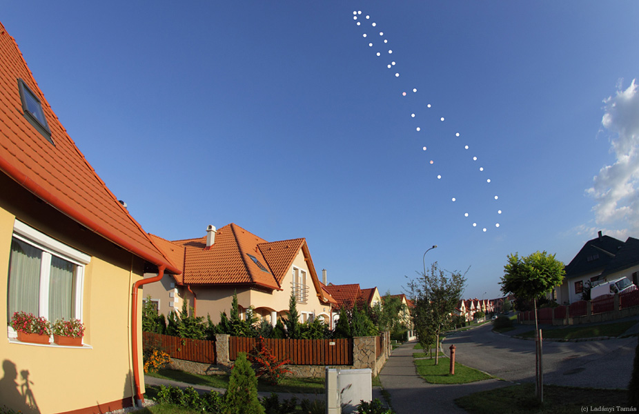 Eine Straße mit Häusern auf der linken Seite, am blauen Himmel darüber ist eine nach links geneigte Achterschleife aus Sonnenbildern, ein sogenanntes Analemma.
