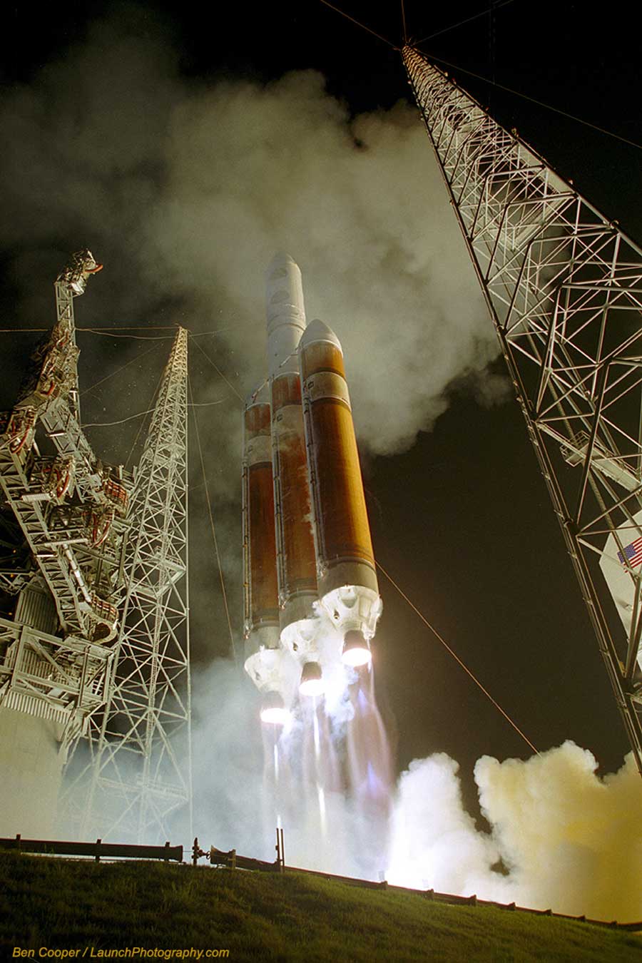 Eine Rakete mit zwei Boostern steigt von der Startrampe auf, umgeben von Rauchwolken und Türmen aus Metallgestänge. es ist Nacht.