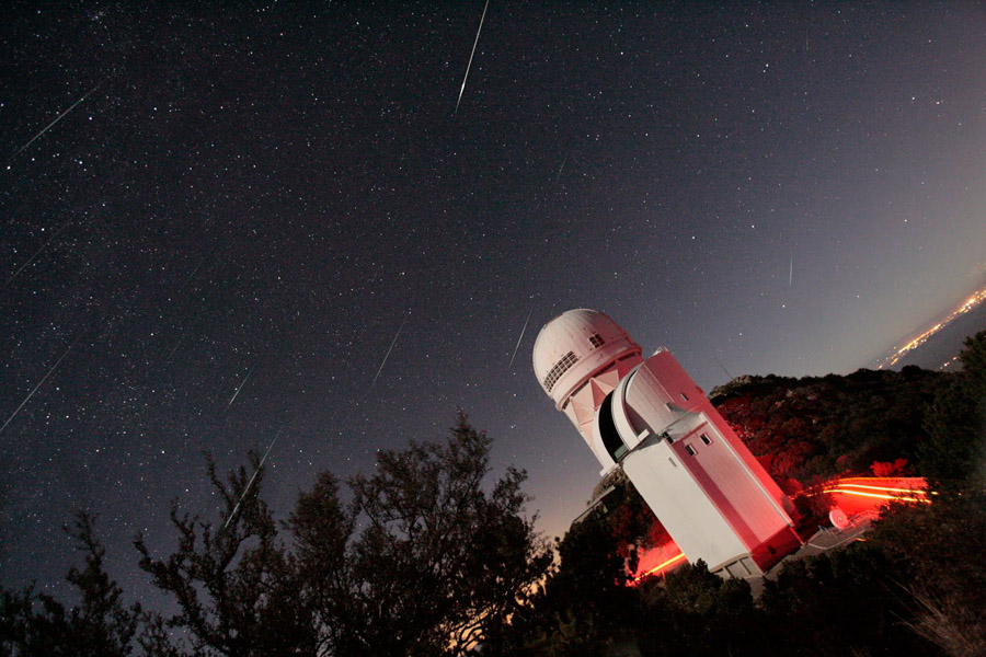 Hinter zwei hell beleuchteten Teleskopkuppeln ist der Himmel sternklar, zwischen den Sternen verlaufen die Spuren von Meteoren.