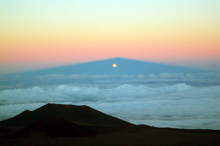 Über ein Wolkenmeer fällt der Schatten eines Vulkangipfels. Unten im Vordergrund sind dunkle Felsen. Über dem Schatten des Gipfels leuchtet der Himmel orangefarben bis hellblau. Unter der Schattenspitze geht der Mond auf.