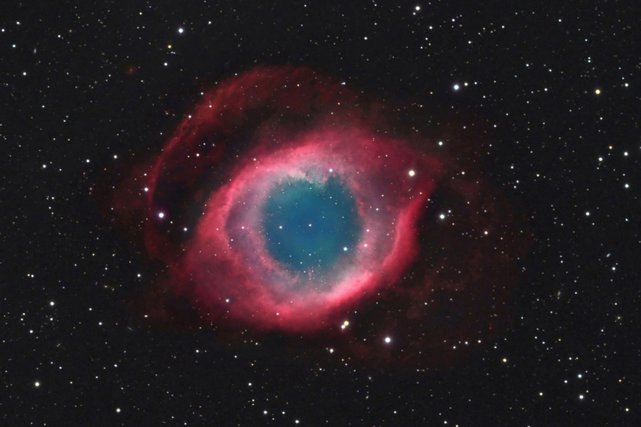 Mitten im Bild leuchtet ein roter Nebelring mit einem zartblauen Inneren, der von wenigen, markant verteilten Sternen umgeben ist.