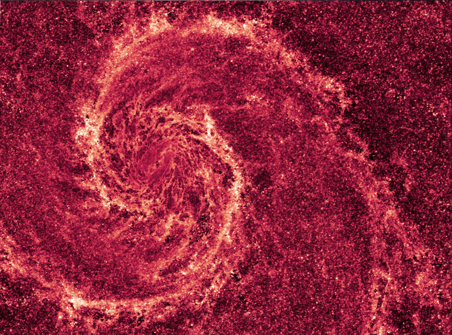 Im Bild breitet sich eine detailreiche, von oben sichtbare Spiralgalaxie aus, mit vielen Staubbahnen, rosa Sternbildungsgebiten und einem hellen gelblichen Zeitrum. Darüber ist eine rötliche Aufnahme in Infrarotlicht gelegt.