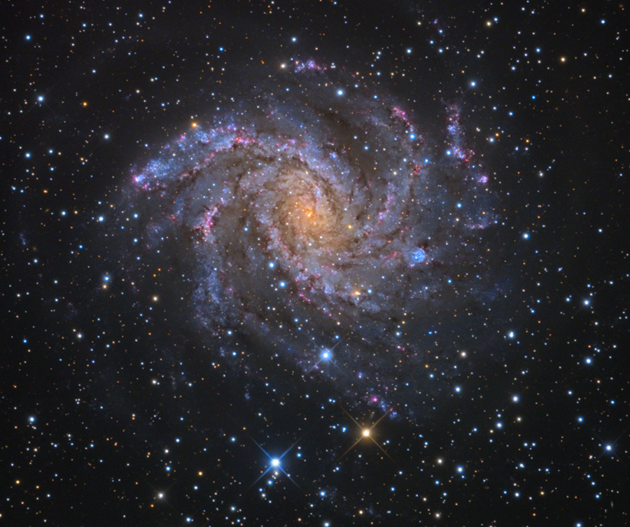 Mitten im sternklaren Bild leuchtet eine von oben sichtbare Süiralgalaxie mit losen Spiralarmen und blauen Sternbildungsregionen. Ihre Erscheinung erinnert an ein Feuerrad oder ein sich drehendes Feuerwerk.