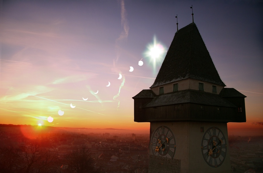 Rechts ragt die Silhouette des Grazer Uhrturms in Österreich auf. Links geht die Sonne schrittweise auf, es sind mehrere Sonnenbilder bei einer partiellen Sonnenfinsternis teilweise vom Mond bedeckt.