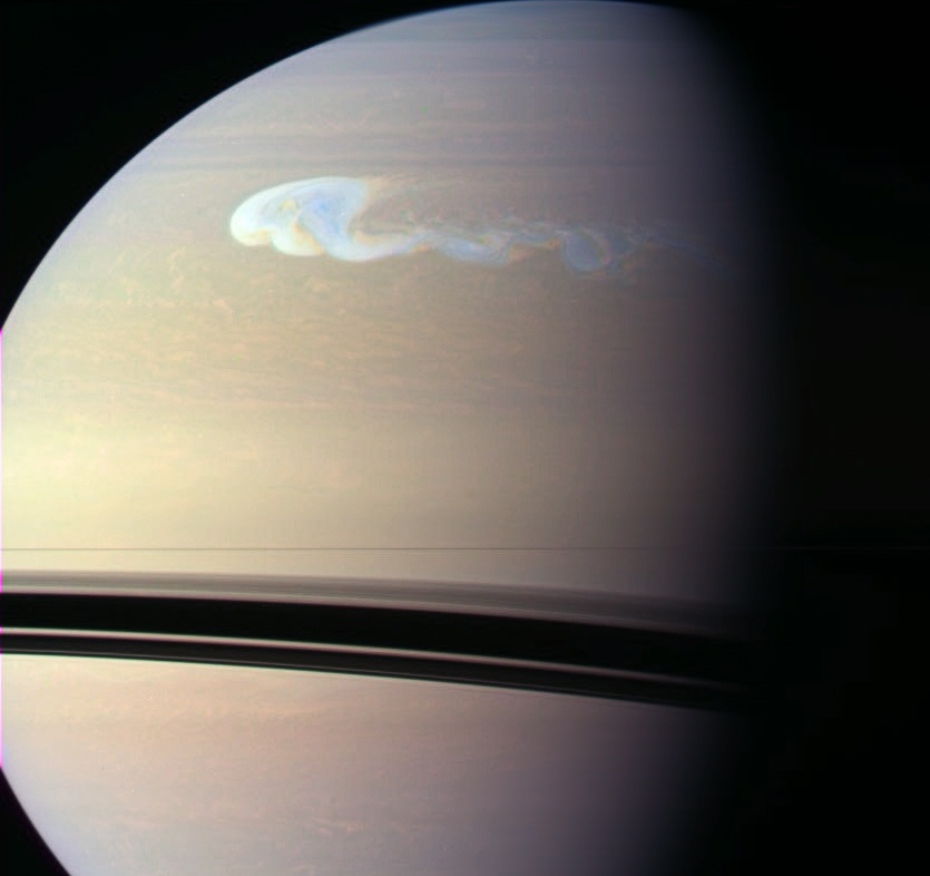 Der Planet Saturn ist nicht vollständig abgebildet, er ragt über das Bild hinaus. Unten sind die Ringe als schmaler Strich abgebildet, darunter die Schatten der Ringe. Oben zeichnet sich auf Saturn ein gewaltiger hellblau-weißer, langgezogener Sturm ab.