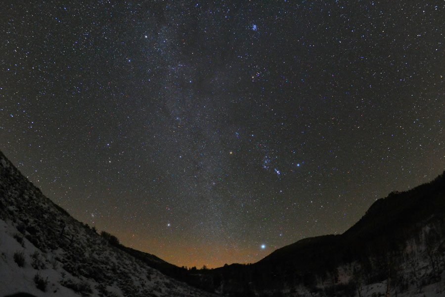 Hinter einer verschneiten Landschaft leuchtet am Horizont schwaches orangefarbenes Licht, von einer Senke in der Mitte steigt die Milchstraße auf, links davon leuchtet das Sternbild Orion, unten der helle Stern Sirius und oben der Sternhaufen der Plejaden. Oben links neben der Milchstraße sind die hellen Sterne Kastor und Pollux.