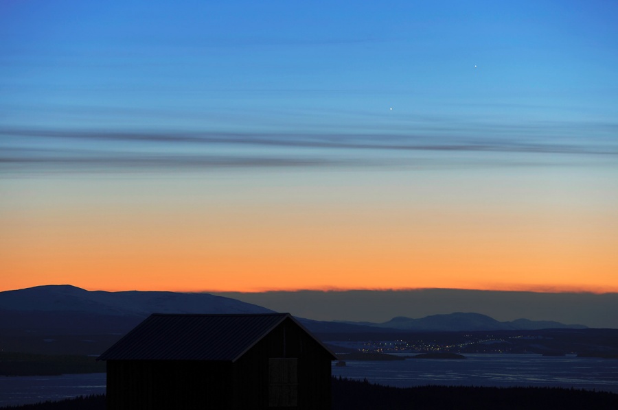 Über der dunklen Landschaft reicht der heitere Himmel mit wenigen Wolkenstreifen von intensiv Orange bis Mittelblau am oberen Rand. Im Westen leuchten zwei Lichtpunkte, Merkur und Jupiter.