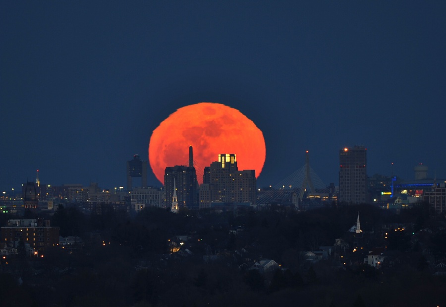 Hinter einer Stadt mit Wolkenkratzern geht der dunkelorange gefärbte Vollmond am nachtbluen Himmel auf. Er wirkt leicht verzerrt und wellig.